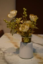 An artificial floral arrangement featuring white roses with gold glitter edges and gold berry sprigs in a white ceramic vase with a metallic gold neck, displayed on a speckled marble tabletop