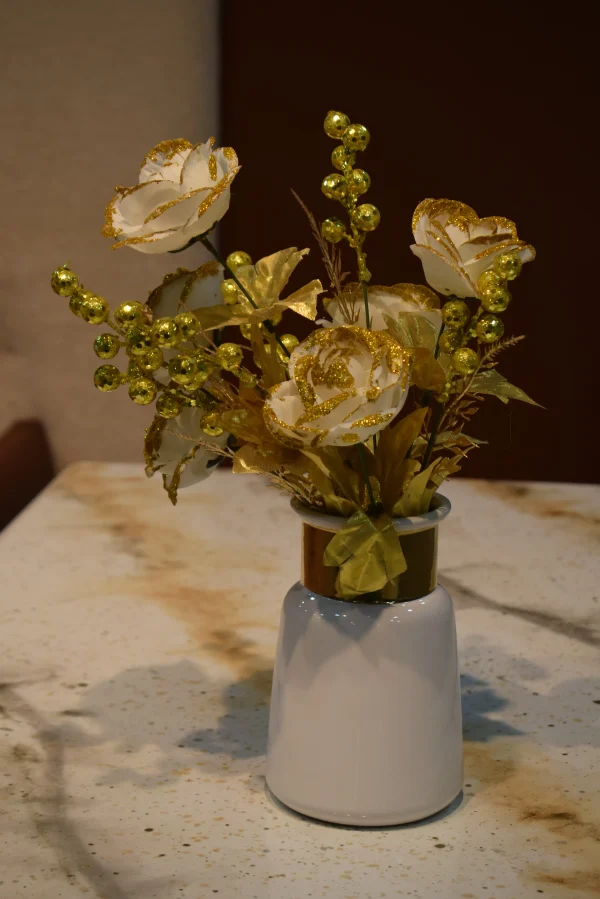 An artificial floral arrangement featuring white roses with gold glitter edges and gold berry sprigs in a white ceramic vase with a metallic gold neck, displayed on a speckled marble tabletop