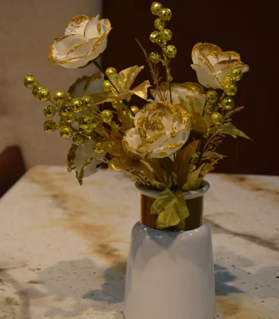 An artificial floral arrangement featuring white roses with gold glitter edges and gold berry sprigs in a white ceramic vase with a metallic gold neck, displayed on a speckled marble tabletop