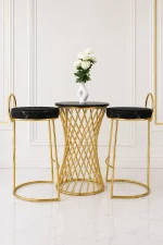 Luxury 3-piece bar bistro set featuring a round black table with a gold woven hourglass base, flanked by two gold bar stools with round black marble-patterned velvet cushions, set against a white paneled wall.