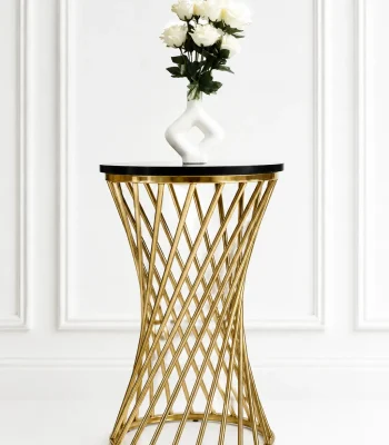 A modern round accent bistro table featuring a polished gold metal hourglass wireframe base and a sleek black tabletop with a white floral vase, set against a white paneled wall.