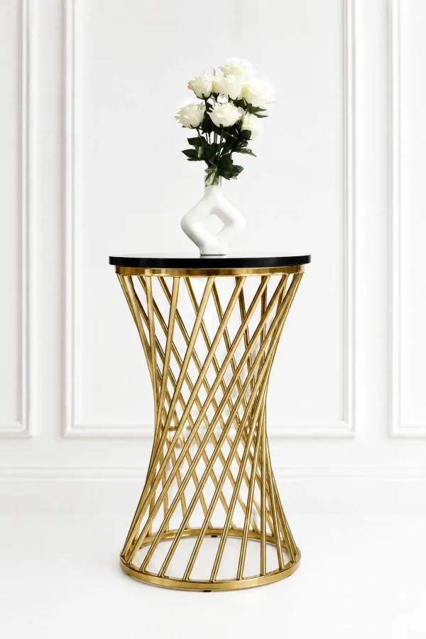 A modern round accent bistro table featuring a polished gold metal hourglass wireframe base and a sleek black tabletop with a white floral vase, set against a white paneled wall.