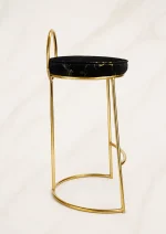 A luxury counter-height bar stool showcasing a slender polished gold metal frame, a low half-moon backrest, and a round black velvet cushion with metallic gold marble veining, against a neutral backdrop.