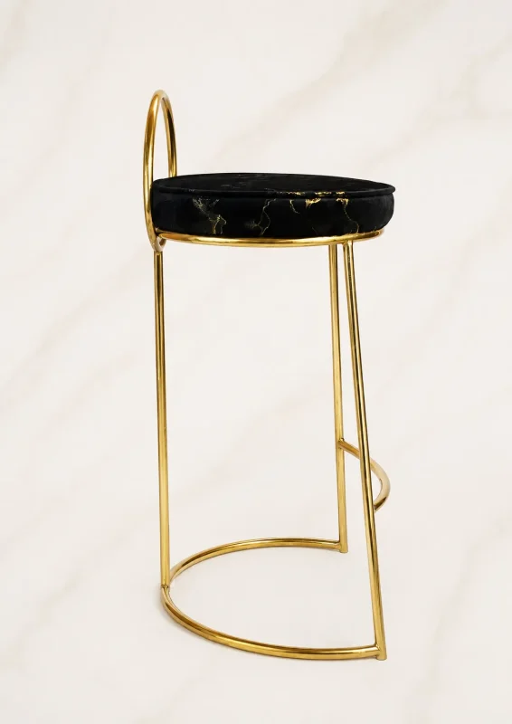 A luxury counter-height bar stool showcasing a slender polished gold metal frame, a low half-moon backrest, and a round black velvet cushion with metallic gold marble veining, against a neutral backdrop.