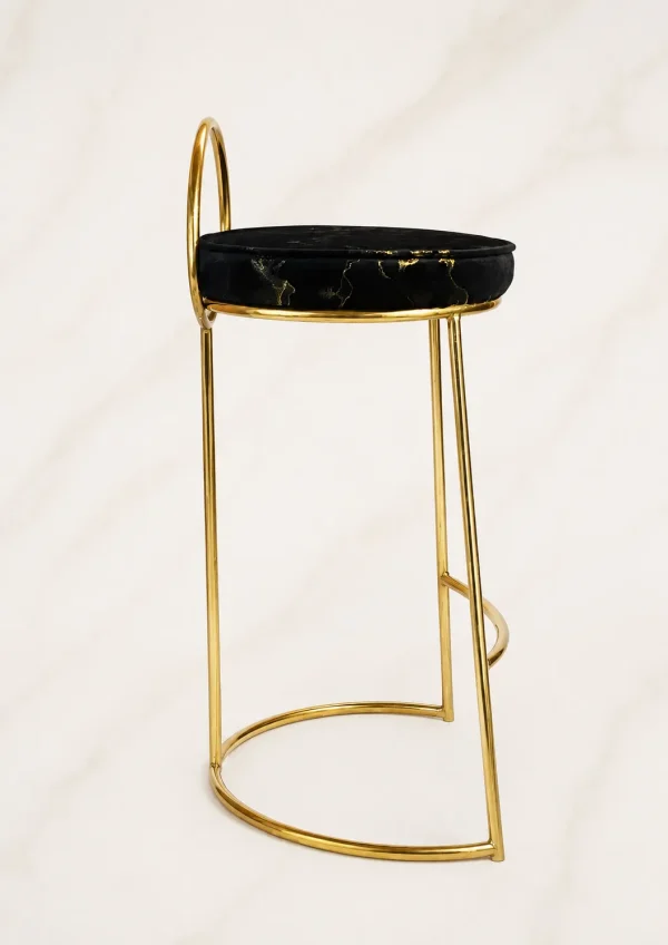 A luxury counter-height bar stool showcasing a slender polished gold metal frame, a low half-moon backrest, and a round black velvet cushion with metallic gold marble veining, against a neutral backdrop.