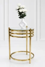 A modern luxury round side table featuring a clear glass top, a polished gold metal frame with a three-tiered stacked ring design under the apron, and a matching circular floor base set against a white wall.