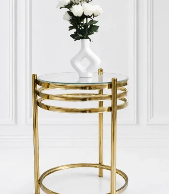 A modern luxury round side table featuring a clear glass top, a polished gold metal frame with a three-tiered stacked ring design under the apron, and a matching circular floor base set against a white wall.