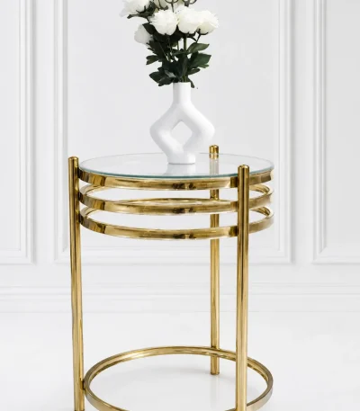 A modern luxury round side table featuring a clear glass top, a polished gold metal frame with a three-tiered stacked ring design under the apron, and a matching circular floor base set against a white wall.