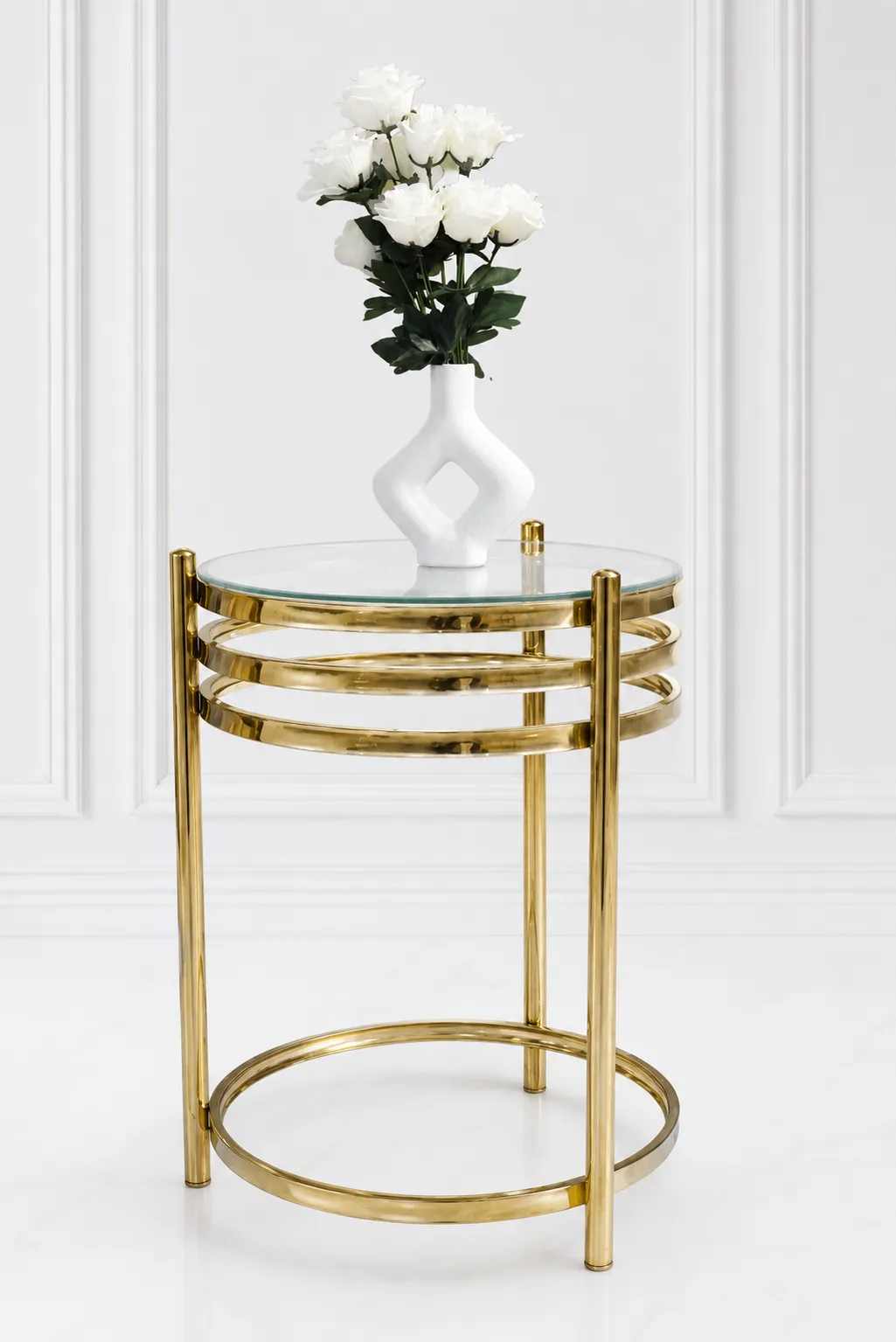 Aura Luxury Round Gold Side Table with Clear Glass Top – Modern Tiered Accent Table A modern luxury round side table featuring a clear glass top, a polished gold metal frame with a three-tiered stacked ring design under the apron, and a matching circular floor base set against a white wall.
