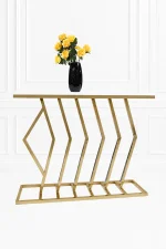 A modern gold console table featuring a geometric chevron-patterned metal frame and a smooth white top surface, styled with a black vase holding yellow roses against a white paneled wall background