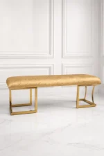 Luxury gold upholstered bench from featuring a textured golden patterned seat cushion and a polished metallic frame against a white paneled wall.