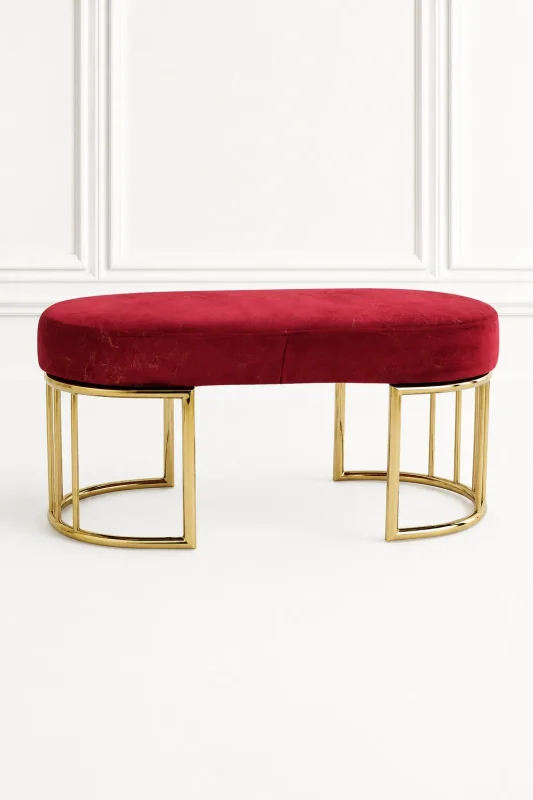 Luxury red velvet gold bench from featuring an oval upholstered crimson seat and polished, semi-circular geometric cage legs against a white wall.