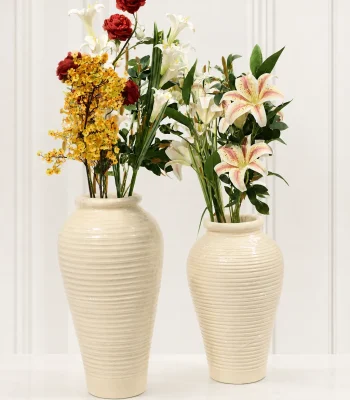 Two tall cream ribbed ceramic vases holding faux white lilies and mixed colorful flowers