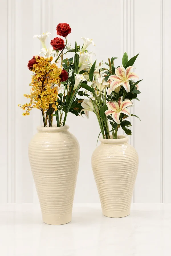 Two tall cream ribbed ceramic vases holding faux white lilies and mixed colorful flowers