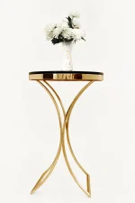 Round gold accent table from featuring an elegant curved pedestal base and a dark circular top supporting a white flower vase.