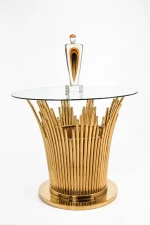 Modern round side table featuring a clear glass top and a pedestal base made of polished gold vertical metal rods, shown with a decorative art piece on top.