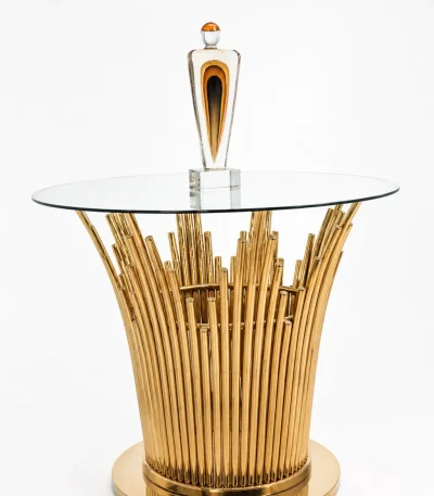 Modern round side table featuring a clear glass top and a pedestal base made of polished gold vertical metal rods, shown with a decorative art piece on top.