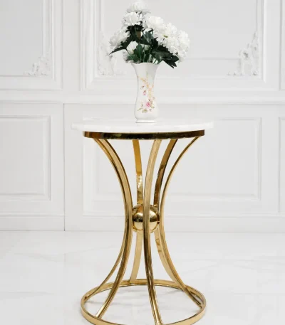 Modern round side table with a polished white marble top and a decorative hourglass-shaped gold metal frame base, holding a white flower vase against a classic paneled wall.