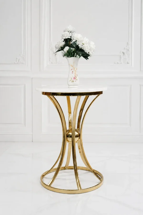 Modern round side table with a polished white marble top and a decorative hourglass-shaped gold metal frame base, holding a white flower vase against a classic paneled wall.