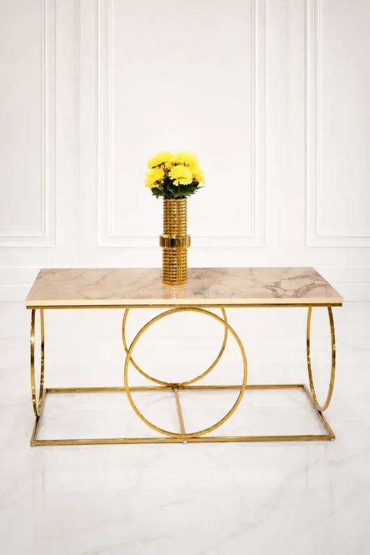 Luxury modern rectangular coffee table featuring a polished beige marble top and a geometric base made of interlocking polished gold metal rings, styled with a gold flower vase.