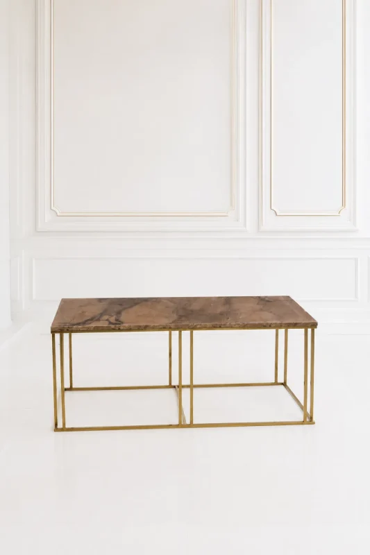 Modern coffee table with a brown veined marble top and a geometric brushed gold metal frame set against a white paneled wall.
