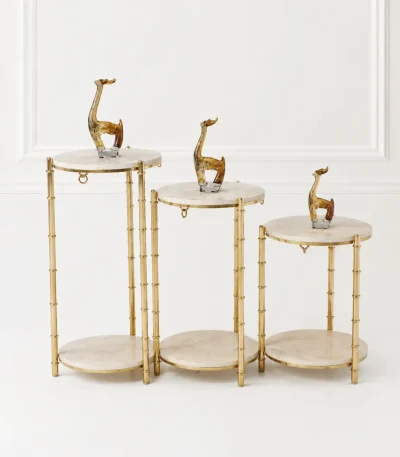 A set of three tiered round pedestal display stands with light beige marble tops and bamboo-textured gold metal frames holding giraffe figurines.