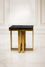 A luxury square side table with a high-gloss black marble-patterned top and a substantial polished gold cross pillar base on a marble floor.