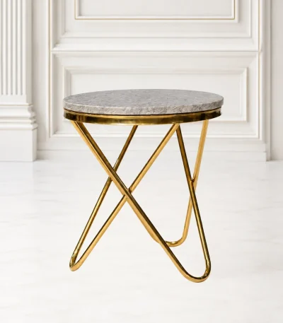 Round gold marble side table with a polished grey stone top and an architectural cross-legged gold metal base, set against a classic white wall panel interior.