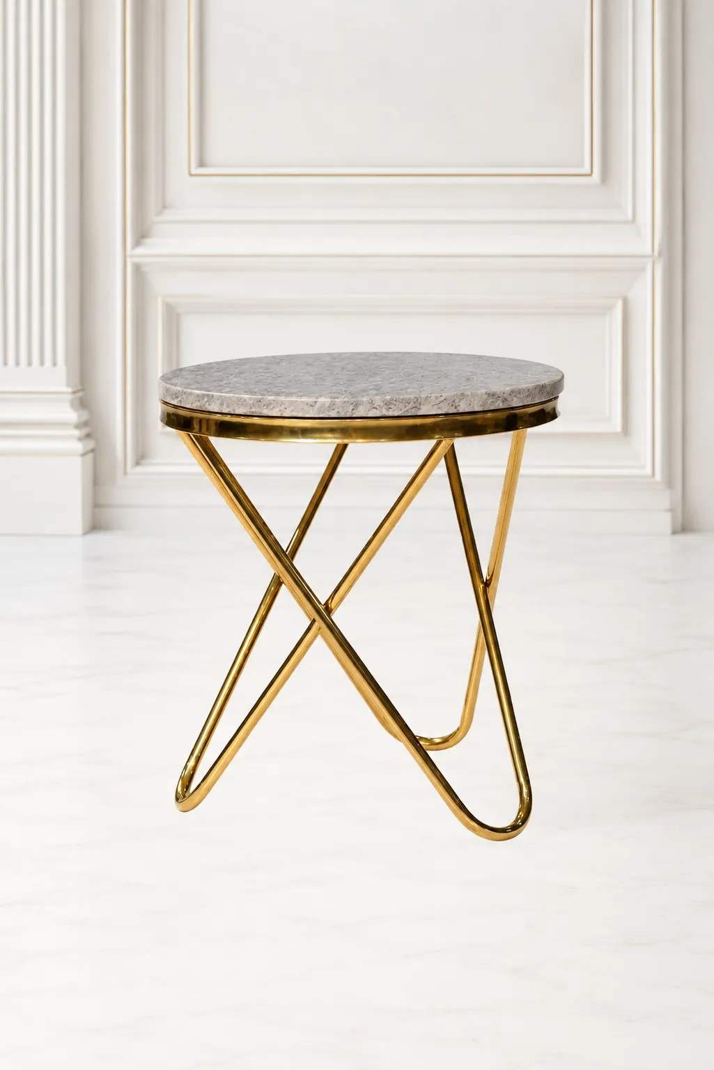 Modern Round Gold Marble Side Table with Sculptural Cross-Legged Geometric Base Round gold marble side table with a polished grey stone top and an architectural cross-legged gold metal base, set against a classic white wall panel interior.