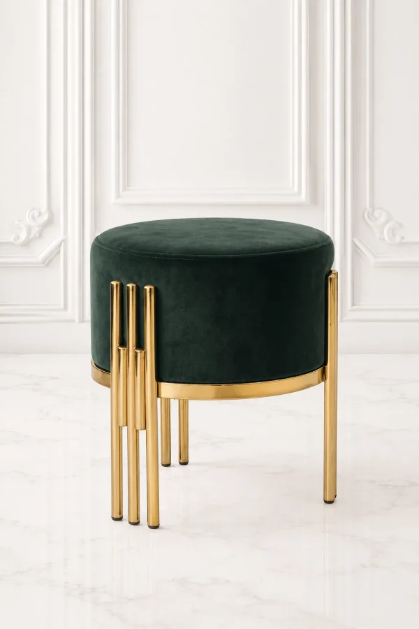 Round emerald green velvet vanity stool featuring a plush cushioned seat and modern tiered gold tubular legs, placed against an elegant white wall paneling background.