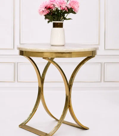 Round cream marble side table with a polished gold crescent-shaped metal base, displaying a white ceramic vase with pink flowers against a white wall molding background