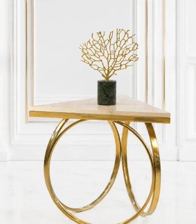 Triangular cream marble side table featuring an interlocking gold ring metal base, displaying a gold coral sculpture on a dark green marble pedestal against a white wall panel background.