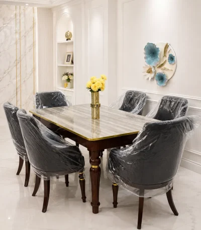 Luxury marble dining table set from featuring a polished gray marble rectangular table with gold trim, dark wood legs, and six matching dark leather tufted chairs in a bright white dining room.