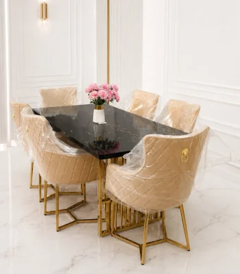 A luxury 6-seater dining set featuring a rectangular black marble tabletop with gold stainless steel legs, styled with six beige tufted velvet chairs under protective plastic wrapping in a bright, modern white dining room.