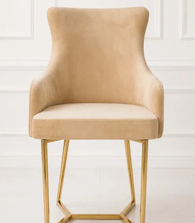 Luxury beige velvet dining armchair with low armrests, supported by a geometric angular gold stainless steel sled base, set against a white paneled wall.