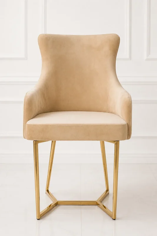 Luxury beige velvet dining armchair with low armrests, supported by a geometric angular gold stainless steel sled base, set against a white paneled wall.