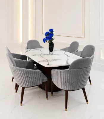 A luxury 6-seater dining set showcasing a white marble race-track tabletop on a dark wood fluted pedestal base, surrounded by six grey channel-tufted velvet barrel chairs with gold-tipped legs in a bright white dining room.