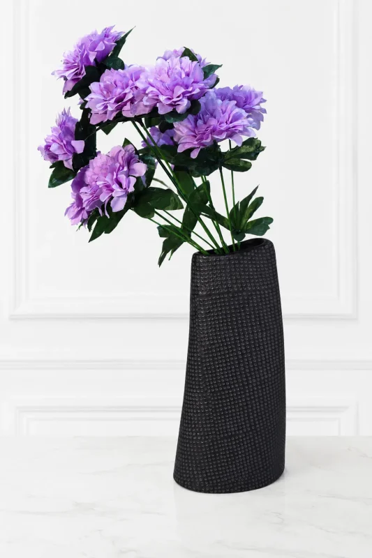 Tall black textured ceramic vase holding artificial purple flowers on a marble surface